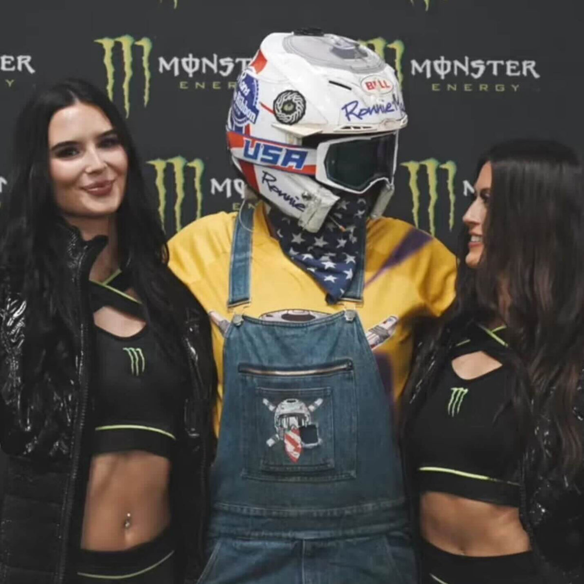 Two women in Monster Energy outfits pose with a man in a helmet and overalls at a promotional event.