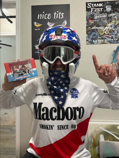 Person in motocross gear holding a Ronnie Mac bike replica, celebrating with a thumbs-up in a cool indoor setting.