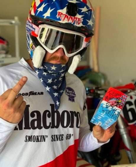 Ronnie Mac showing off a signed item, wearing a patriotic motocross outfit and helmet in a garage setting.