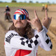 Man with mullet, wearing American flag bandana and sunglasses, making rock hand gesture at motocross track.