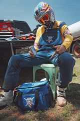 Rider in blue overalls sits with a Ronnie Mac Soft Cooler on a chair, showcasing the crossbones logo outdoors.